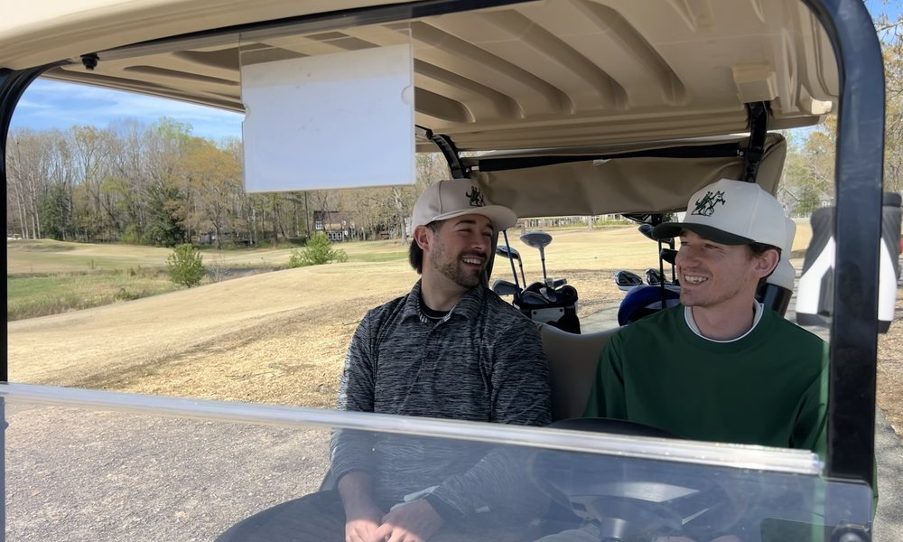 Two golfers in Shaggy Golf hats riding a golf cart on the course